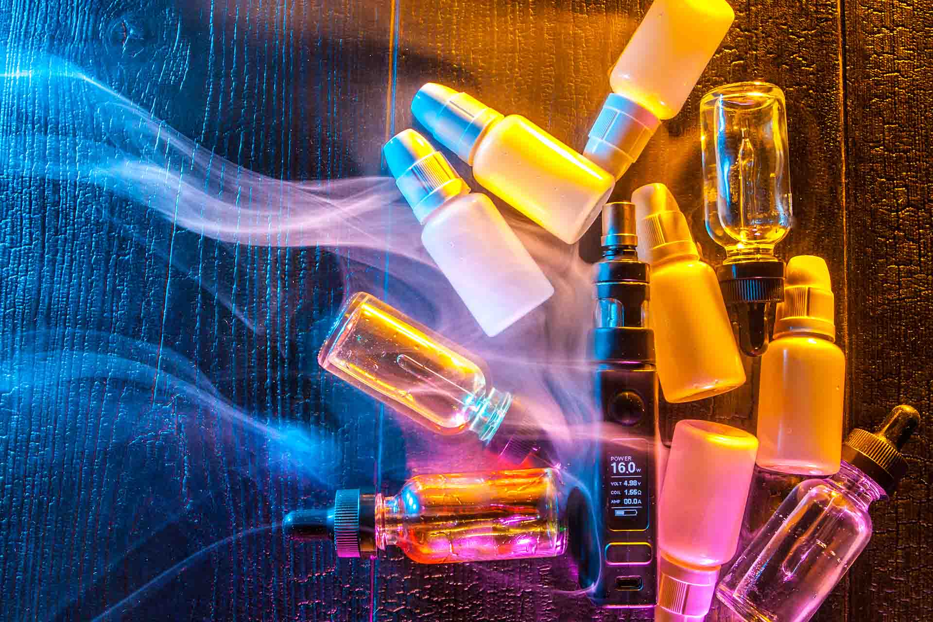 Bottles of e-liquid on a table with vape, surrounded by vapour (Image)
